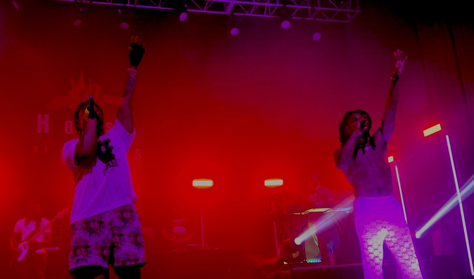 WowGr8 (left) and Olu (right) lift their hands towards the crowd while performing song “STRONG FRIENDS”. Red and purple lights illuminate the stage behind them. The House of Blues logo, which is a heart on fire with the words “House of Blues” across it, can faintly be seen behind them along with their bassist, DJ, and keyboarder.