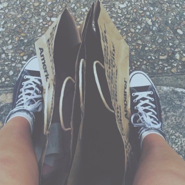 Black converse shoes with two paper American Apparel bags between the two shoes. The bag and shoes are on concrete.