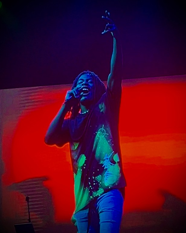 PlayThatBoyZai is illuminated by blue light while he raises his hand towards the crowd. He is wearing a black graphic t-shirt and skinny jeans. He is wearing a gold grill set in his mouth and has a chain around his neck. The background is a red screen. 