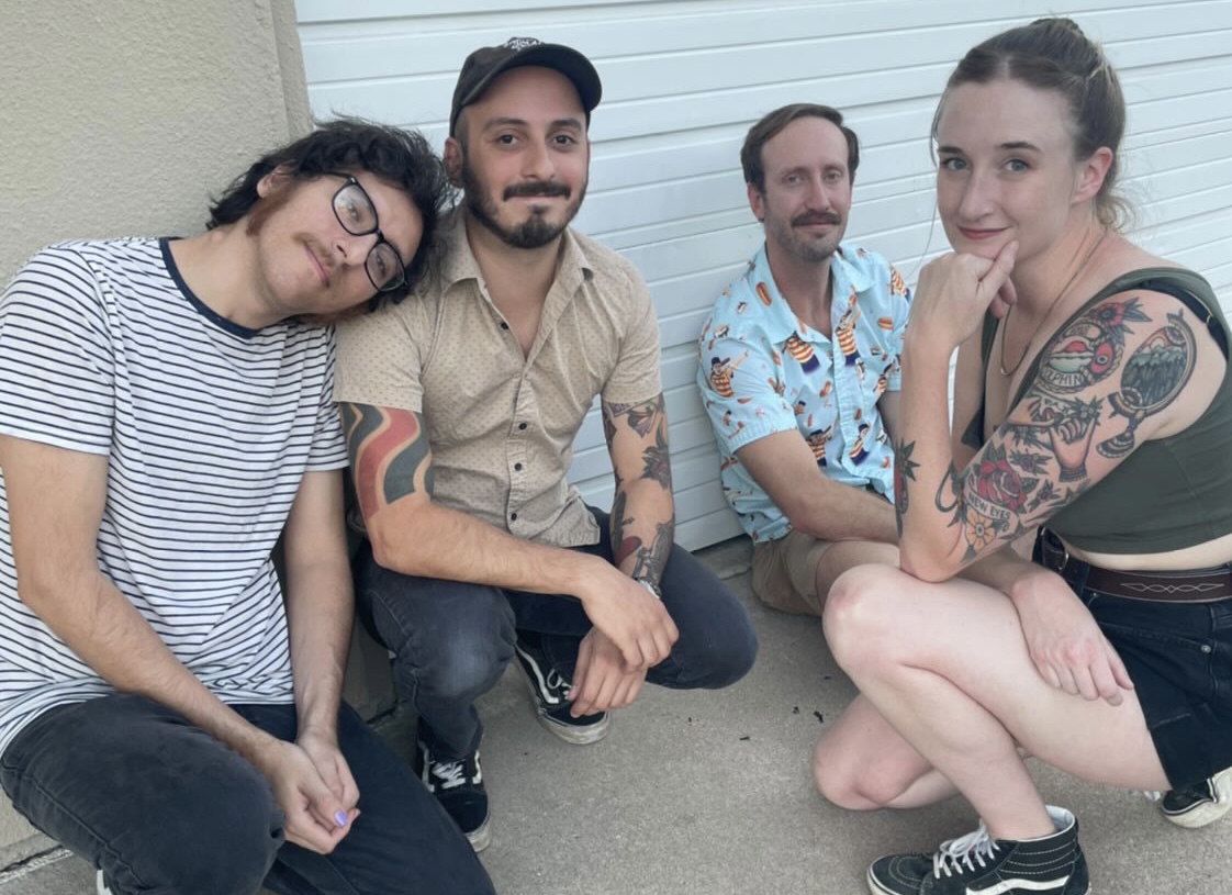 Photograph depicts the four members of Lola Tried, crouched down in a casual position on top of what looks to be a concrete sidewalk. Band Member farthest to the left is pictured with shaggy hair, a mustache, a black and white striped t-shirt and black jeans. Member depicted in middle left of the photo wears a black baseball cap, a beige button down shirt, black jeans and black Vans sneakers. Member depicted in middle right of the photo is partly hidden by member on far right but can be seen wearing a brightly colored, yellow and aqua button down shirt with khaki shorts. Member on far right is shown with her right arm propping up her chin, showcasing her half sleeve of colorful tattoos; she is wearing a green cropped shirt, denim shorts and black hi-top Vans sneakers.