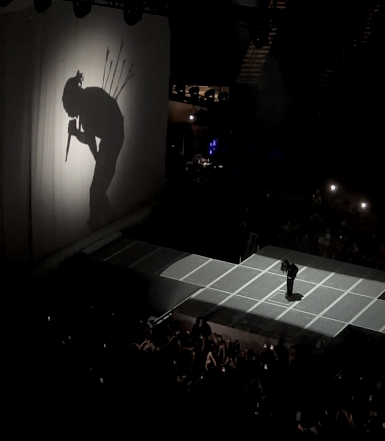 Kendrick Lamar is crouched over singing towards the crowd, holding a microphone to his mouth. A spotlight beams on him, and a large-scale silhouette can be seen behind him projected on long white drapes. On Lamar’s shadow of his crouched figure holding a microphone, arrows can be seen stabbing him in the back