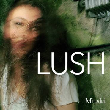 The album cove is an image of mitski in front of a mossy stone wall with the words Lush in all caps , and her face is blured as if it was taken in motion of her shaking her head.