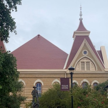 A picture of Old Main as seen from a distance. To the left, there are tree crowns and in front is the vaquero statue and a lamp post with a texas state banner.