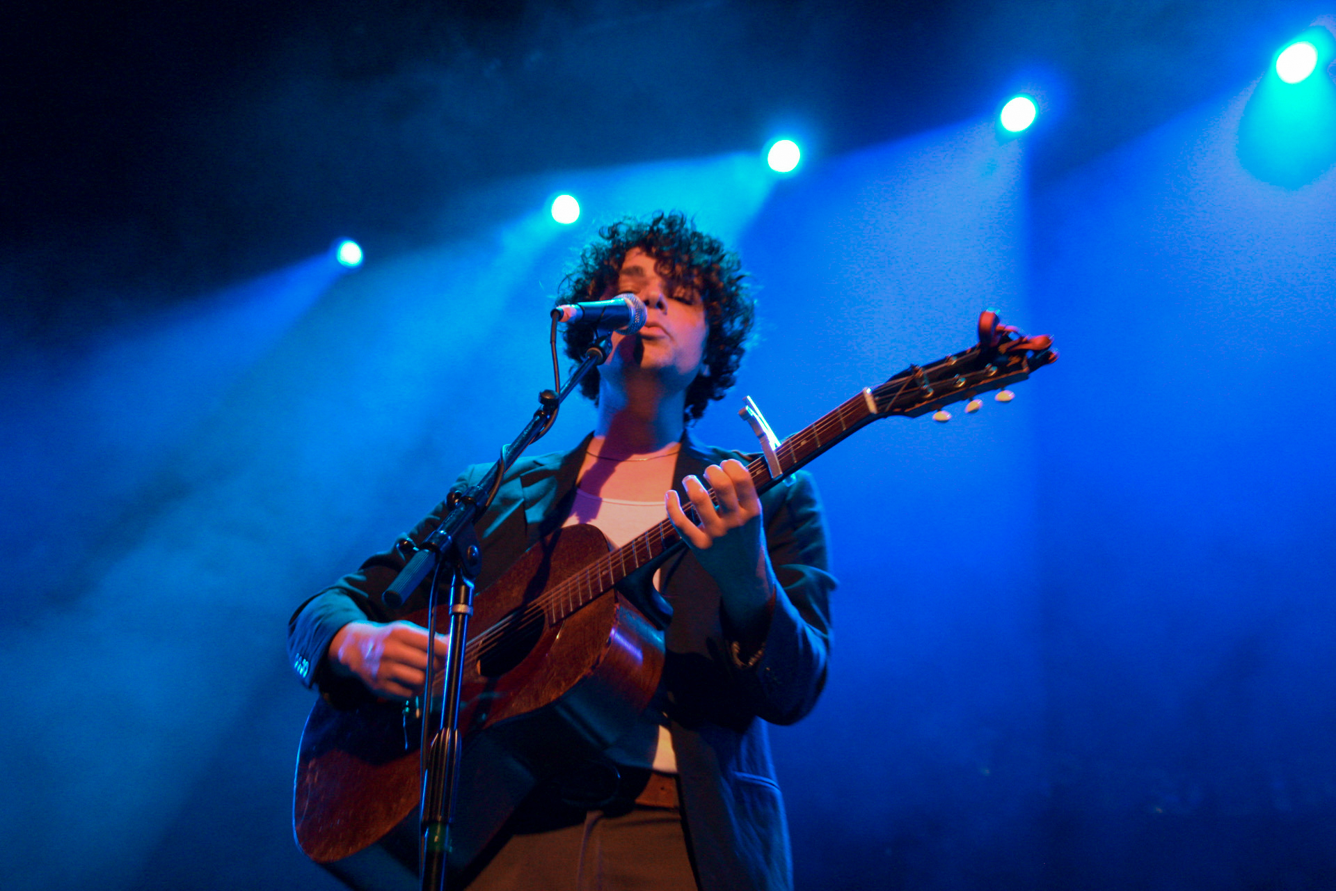  Charlie Hickey playing acoustic guitar under blue lighting in a dark suit jacket atop a white shirt. 