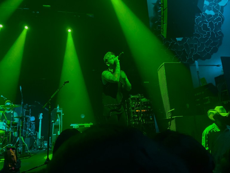 : A man with two colored hair, sings into the microphone on stage under a green light