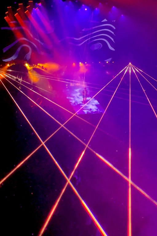 The photo is mainly illuminated by purple and pink lights while orange laser beams can be seen beaming over the crowd, intersecting each other. Cudi can be seen in the middle of the stage standing on top of the stage that is displaying clouds of smoke underneath his feet. A display screen behind Cudi shows a swirling black-and-white image. Cudi is wearing his custom blue Givenchy sweater that has his screaming face with his hands on his cheeks on the back of it. 