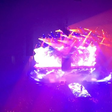 The majority of the photo is illuminated with red lights while orange strobe lights can be seen flared above the crowd. Cudi can be seen with one hand raised, and the other hand holding his microphone up to his mouth on the big display screens at the top left and right corners of the picture. The audience’s silhouette can be seen at the bottom perimeter of the picture. The mixing booth can also be seen towards the bottom half of the picture.