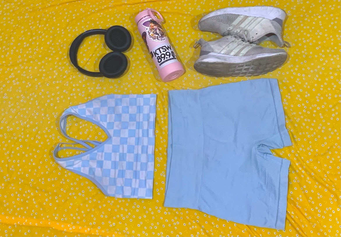 The photo features a yellow background with small black and white flowers. On top of the fabric, there is a workout set of blue biker shorts and a blue and white checkered sports bra. Next to the shorts, there is a pair of black headphones with a pink water bottle beneath it. The water bottle has a KTSW 89.9 logo sticker on it and a sticker of a witch holding a cat above it. Beneath the water bottle, there is a pair of white and gray Adidas sneakers.