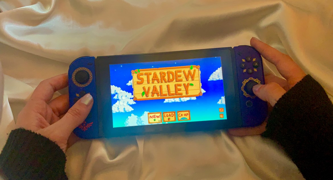 The photo features a white silk background. There are two hands holding a Nintendo Switch. The switch has blue and purple joycons, and the screen shows the title screen for Stardew Valley. There are green trees behind the brown lettering.