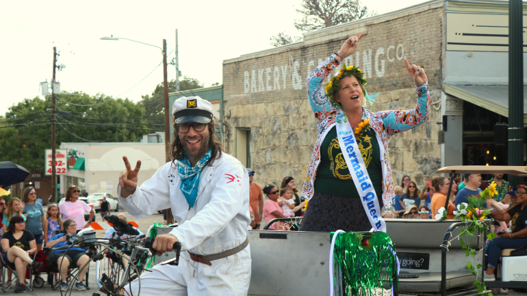 A pedicab drives a woman standing up in the carriage. The driver is wearing a white jumpsuit and sailor hat and is posing for the camera with a peace sign. The woman in the cab has her hands raised wearing a white sash that reads “2022 Mermaid Queen”. The pedicab is decorating with vines and yellow flowers. 