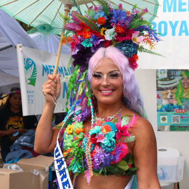 A woman dressed as a mermaid poses for the camera holding a teal parasol above her head. She is wearing a multi-colored floral headpiece; a matching multicolored top; a pink, purple and green ombre wig; a multicolored mermaid necklace and colorful statement makeup. She is standing in front of the Mermaid Society of Texas booth at the Downtown Street Faire.
