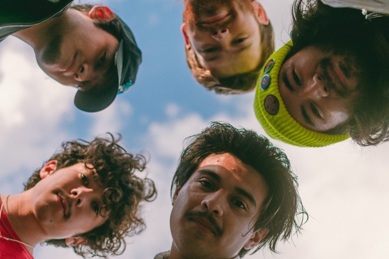 Headshot of the band Flight by Nothing. They are standing in a circle with a blue cloudy sky behind them, looking down at the camera.