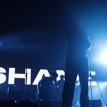 Band Shame positioned in front of a screen reading “SHAME” with blue lighting.