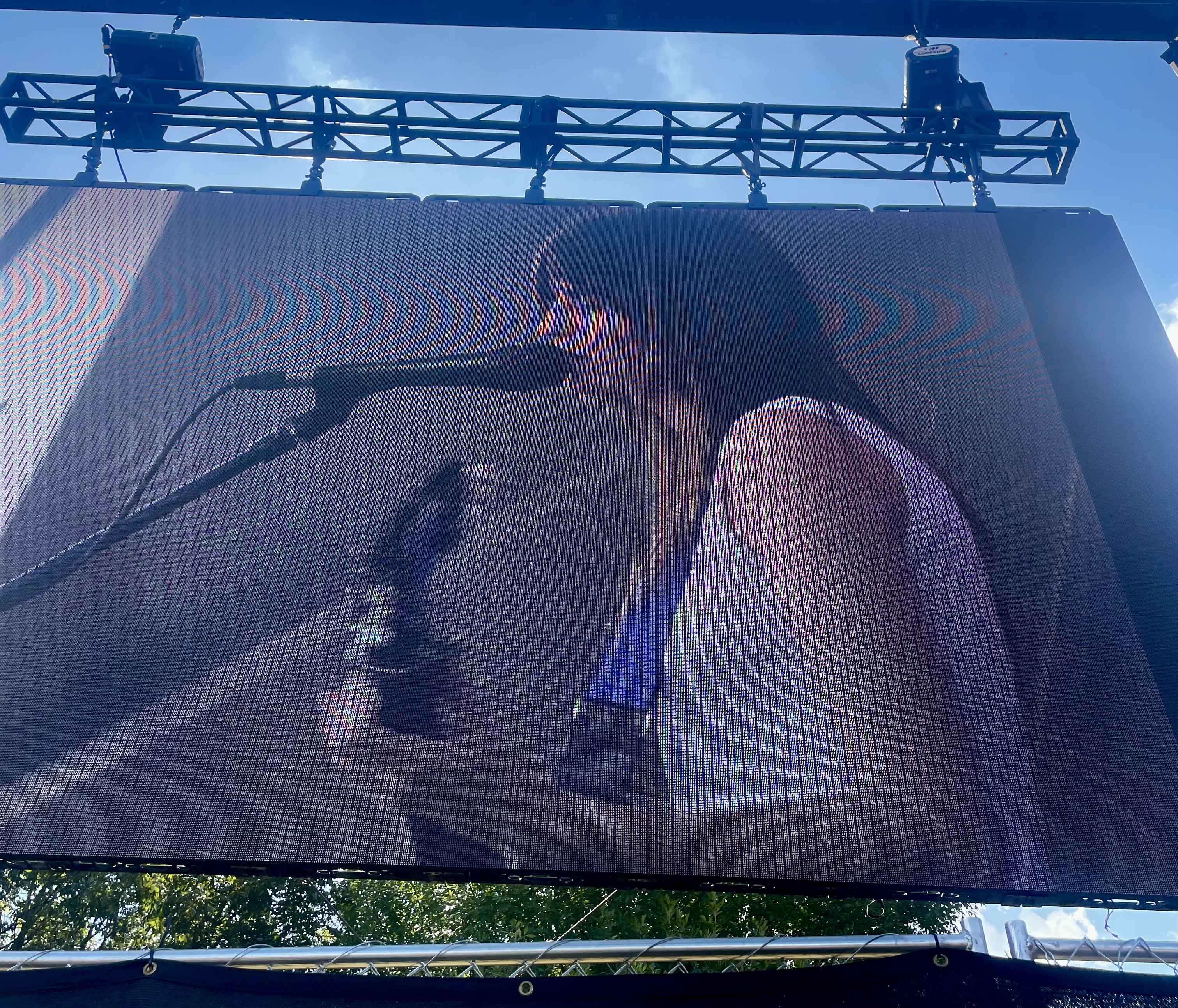 The image features Faye Webster on screen at the Barton Springs stage. She is singing into the microphone while playing the guitar. The screen is slightly distorted from the camera lens. 