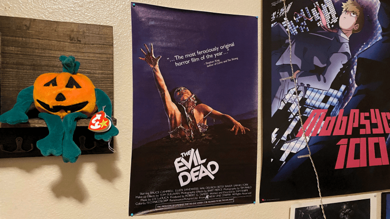 An “Evil Dead” (1981) poster hung on a bedroom wall. The poster depicts a woman reaching for air as she is being dragged underground by a decaying hand. A review for “Evil Dead” reads, “the most ferociously original horror film of the year”. To the right of the “Evil Dead” poster is an anime poster and to the left is a wooden shelf with a Beanie Baby jack-o-lantern sitting on the ledge. 