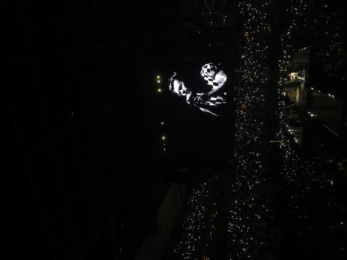 Harry Styles singing sign of the times at the Moody Theater. He is on a black and white screen. Fans use their phone flashlights to light the arena.