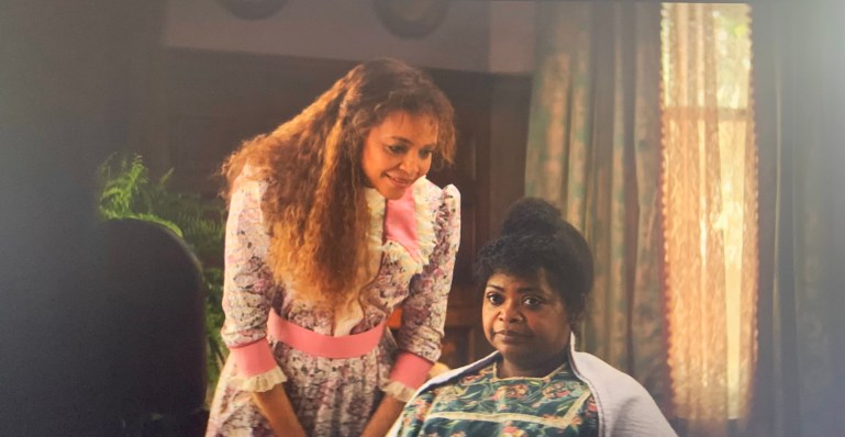 Two women are shown. One is sitting while the other is standing. The woman that’s standing is smiling as she leans over the woman sitting down. The woman sitting down is frowning, clearly displeased by what the girl standing over her is saying.
