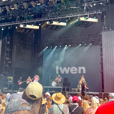 Twen at Austin City Limits 2022 ALT-TEXT: The image features the members of Twen performing onstage. The screen behind them is black with the name twen in gray. The backs of various people’s heads are seen in the forefront.