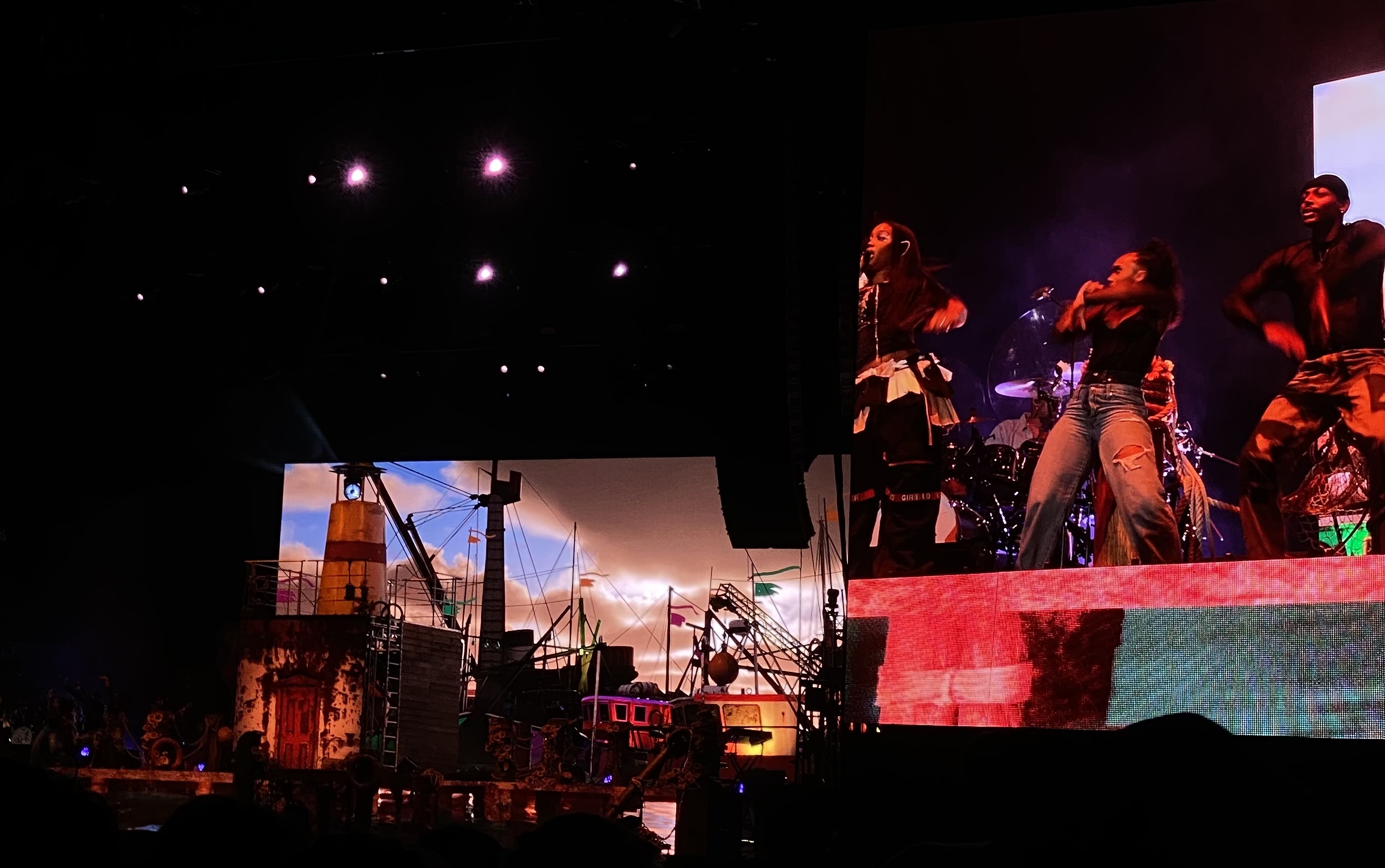 : Image features a close-up view of SZA and two dancers projected on side-stage viewing screen to the right of the stage. An intricate and nautical stage design is seen to the left of the viewing screen, ultimately enhanced by moody, dramatic lighting .