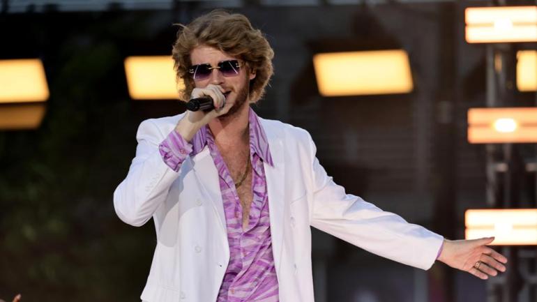 A young man with blonde curly hair holding up a microphone with a white and purple tuxedo