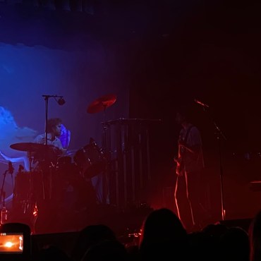 Image features a dimly lit stage with visuals of tropical birds behind the drummer. Farthest to the left, an outline of Giannascoli’s bass player is visible beside an outline of his drummer to the right. In the image’s far right corner, Giannascoli is seen sitting at a piano.