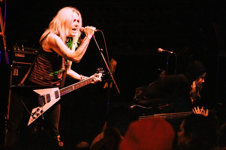 Image features L7 guitarist Donita Sparks with a black and white guitar singing into Mohawk microphone. Sparks is seen with Blonde hair, black tank top, grabbing the mic with her right hand. 