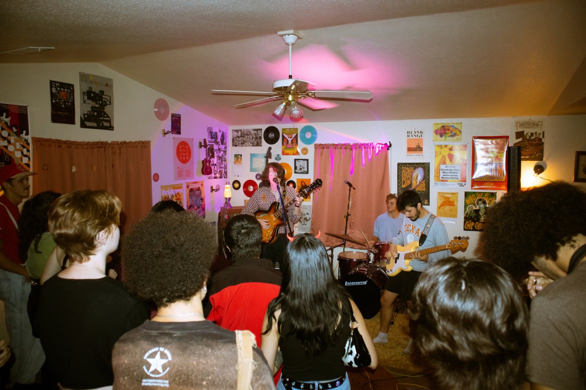 Image features Arbordae (CURTIS ROWE, ETHAN LUGBAUER, CALEB ALVAREZ, RESPECTIVELY) in the middle of performing their Halloween set in the living room of Earle’s Basement. Rowe can be seen singing while playing guitar. Similarly, Lugbauer is seen playing the drums while Alvarez looks down at his yellow bass. A variety of posters fill the walls of the spacious living room while purple lighting is projected from the crowd onto the band. 9 audience members are visible, scattered in the image. DESCRIPTION: Arbordae onstage at Earle’s Basement on November 4, 2022, in San Marcos, Texas.
