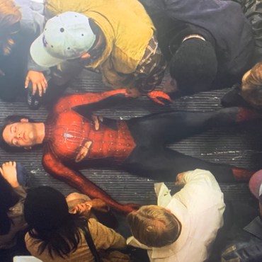 On the ground of a Subway train, lays an unconscious man known as Spider-Man, a man in a ripped red suit, with his mask off while surrounded by lots of people.