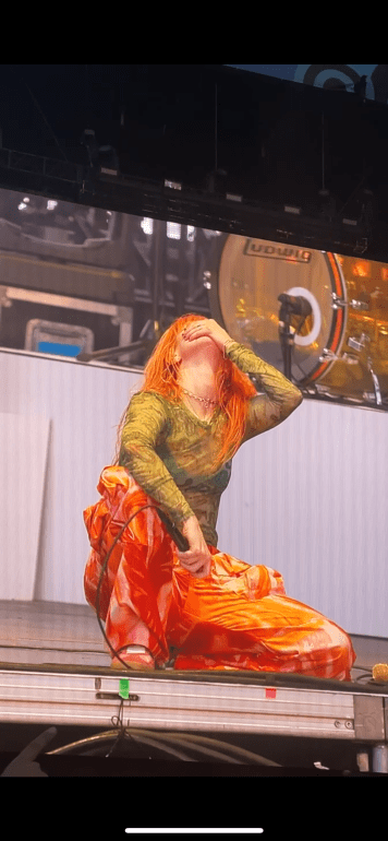 On the screen is Hayley Williams crouched on the floor of the stage with her left hand over her mouth as she looks upward. She is in a green mesh top and orange tie dye pants. 