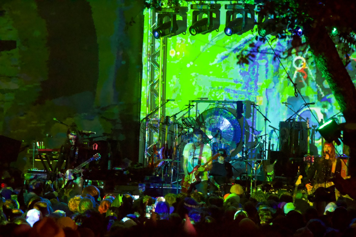 he image features Gaz Liddiard of Tropical F**k Storm on stage playing guitar in the center. Surrounding him are other members of the band, with Erica Dunn on the left, Lauren Hammel behind, and Fiona Kitschin on the right. The audience can be seen in the forefront. Psychedelic visuals are being projected onto the back of the stage.