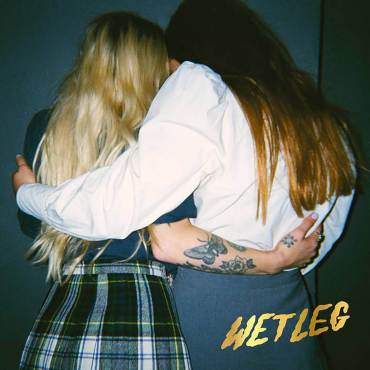 two women are seen embracing from the back. The brunette One is wearing a white shirt with a grey skirt and the other blonde one is wearing a plaid skirt and a navy shirt. The name wet leg is seen printed in the bottom right corner in gold.