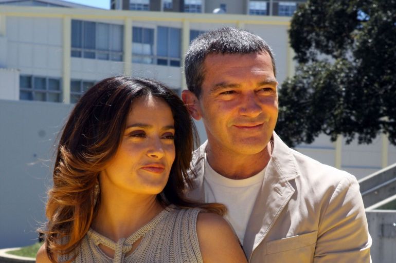 Antonio Banderas and Salma Hayek posing together. He is close behind her to the right from the viewer’s perspective. He is wearing a tan suit jacket and she is wearing a tan dress adorned with belts and ropes.