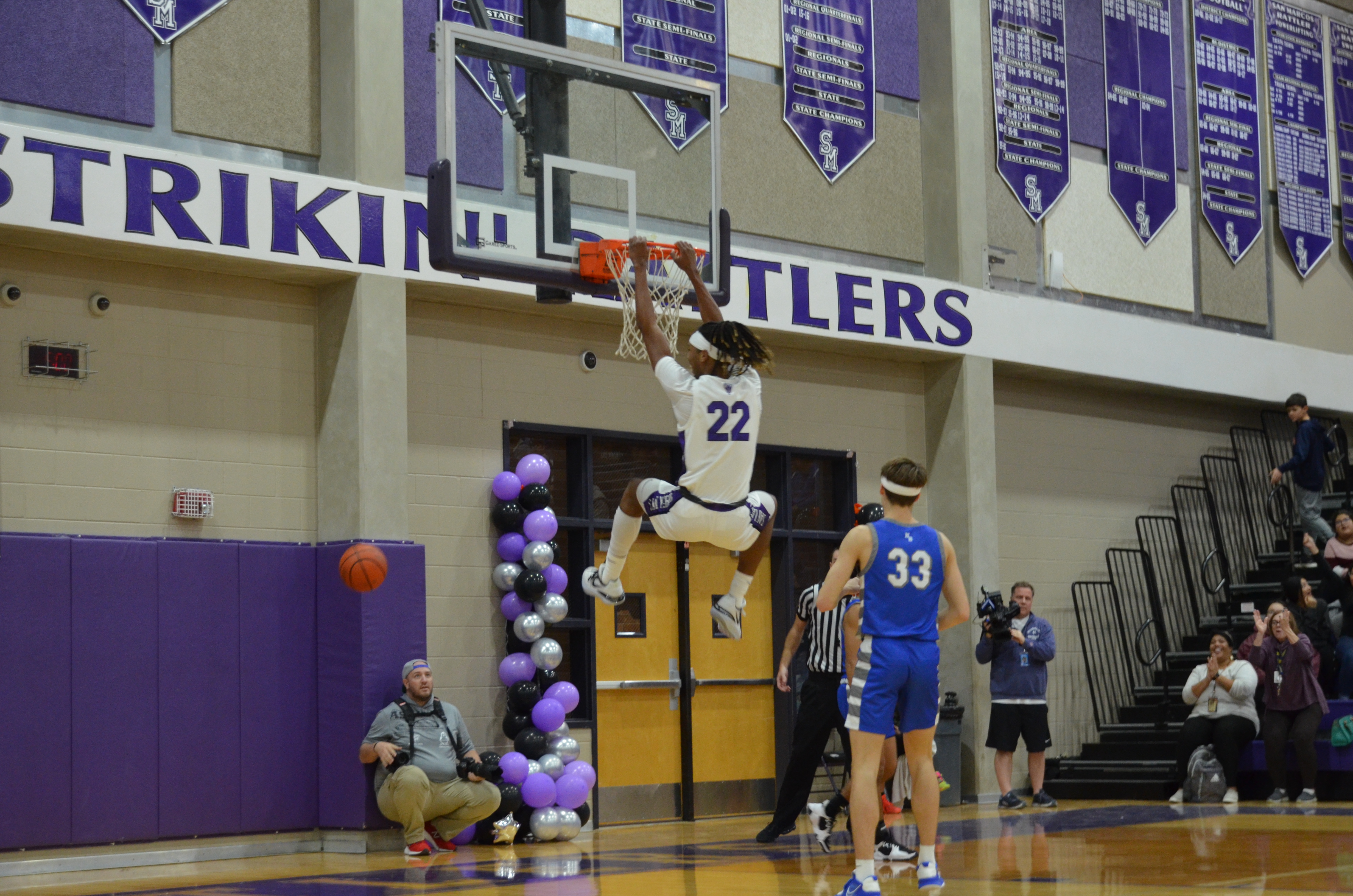 One blue basketball player stands idly on defense while white and purple player hangs from the rim off a big dunk .