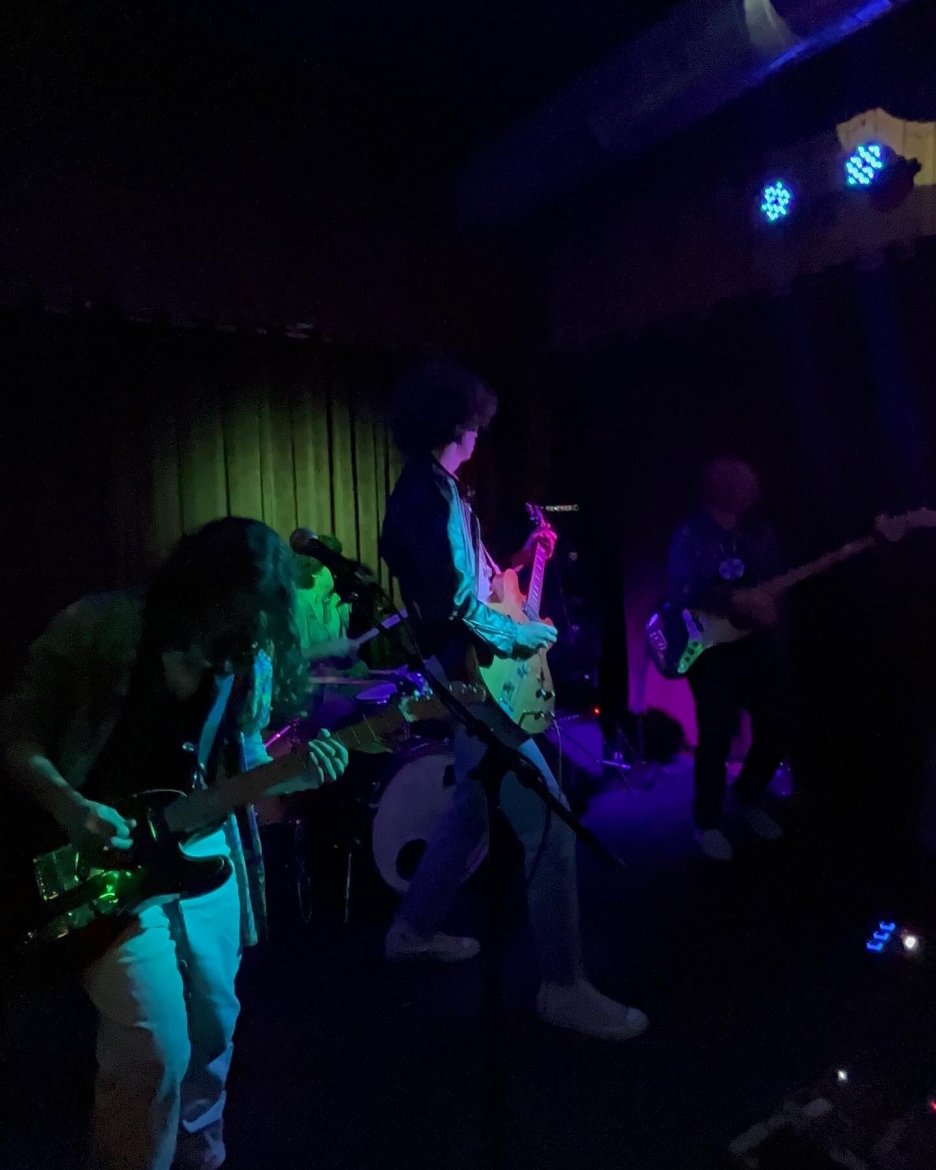 low lighting with only glimpses of green and purple, 3 guys playing guitar, one guy playing drums