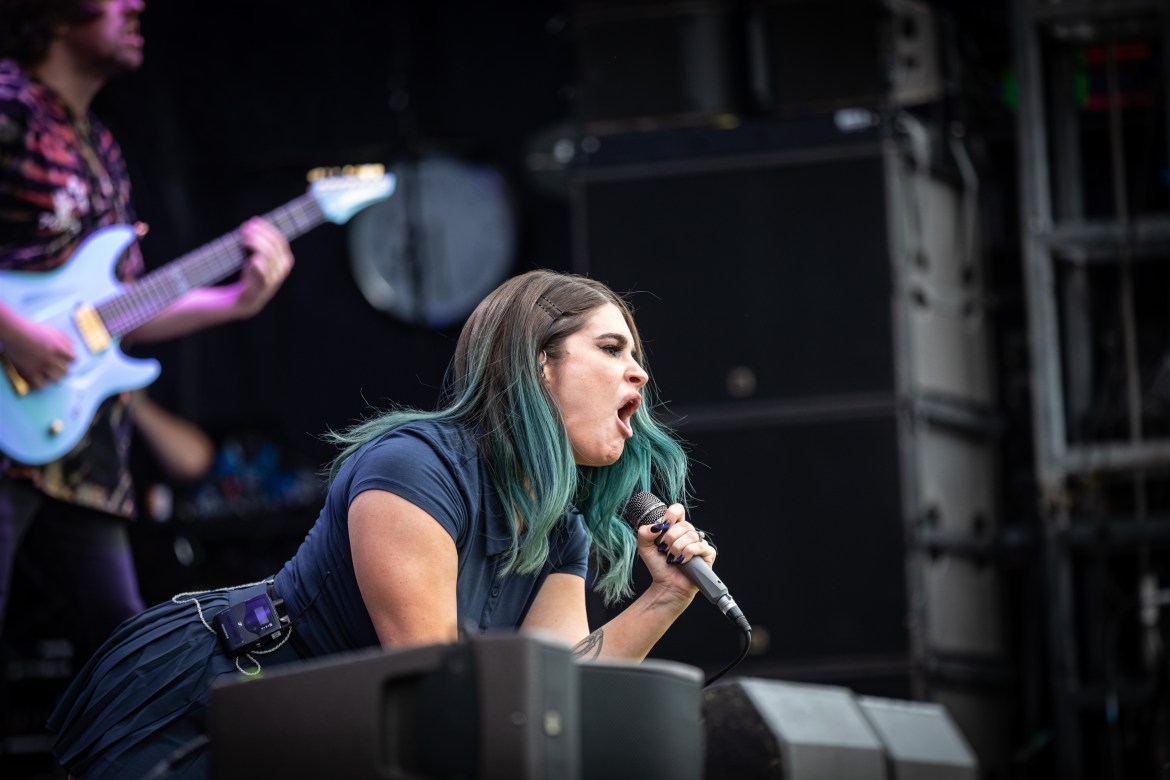 A blue-haired woman is screaming into a microphone, kneeling on the edge of a stage. A man playing a blue guitar is blurred in the top left corner.