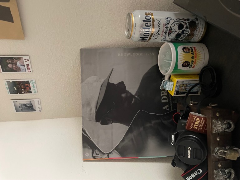 A record sitting on a table behind a mug and camera. The record has a photo of a man with the title of the album “1988” and artist name “Knxwledge.” 