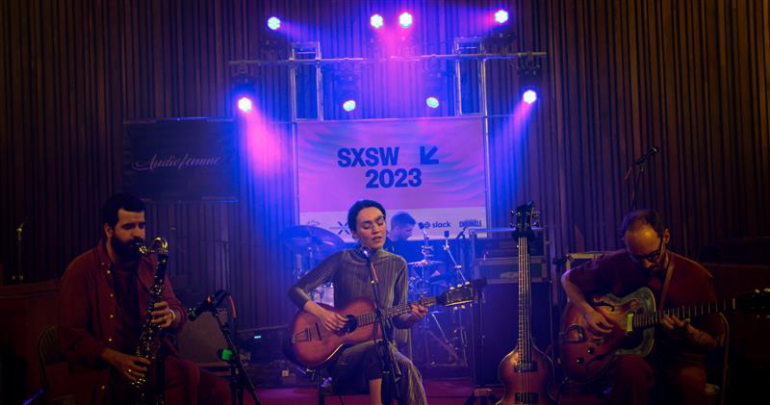 : Núriah Graham plays a guitar centerstage. She is accompanied by a saxophonist and a bassist who sit by her side. The stage is lit with purple lights and there is a banner that reads SXSW 2023 in the back.