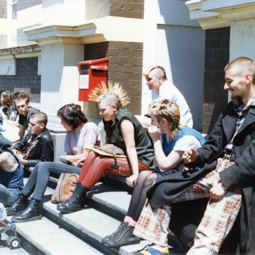 A group of young men and women dressed in dark colors with unusual hair styles siting on stairs outside of a post office in New Zealand in broad daylight.