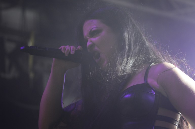 A dark-haired woman makes an angry face into a microphone while screaming with a purple light behind her.