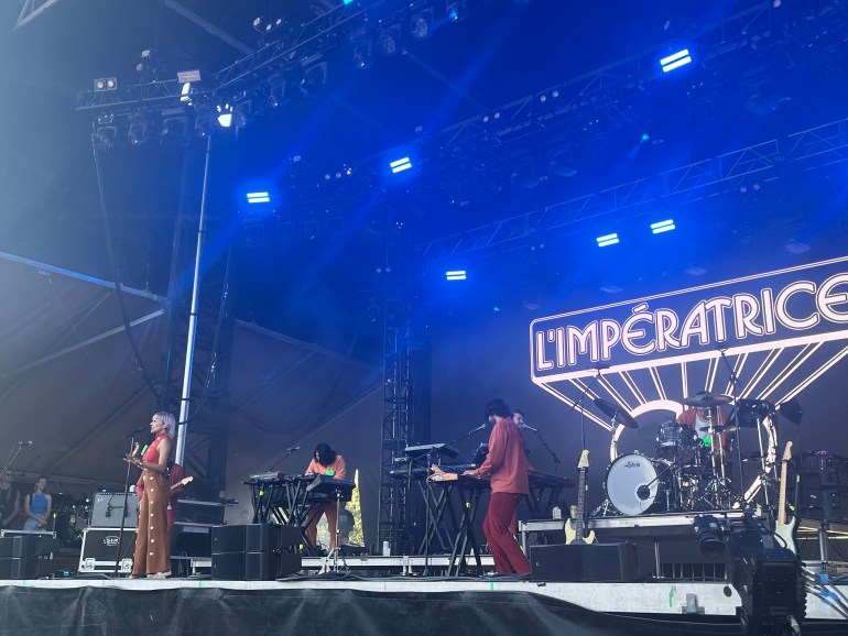The picture displays a stage with the words “L’imperatrice” on a back screen. There are multiple pianists and a lead singer in the picture