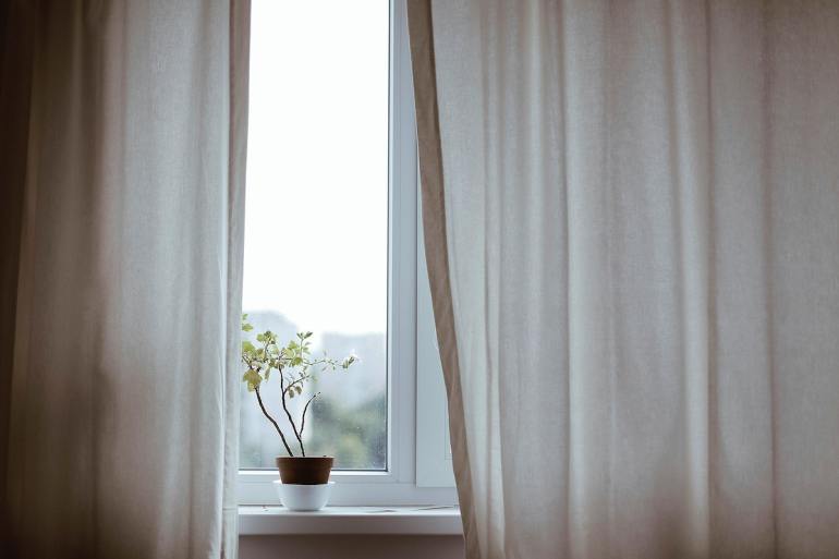 A small potted plant sits on a windowsill. Surrounding it are a set of flowing white drapes. They obscure much of the window in a rather formless fashion.