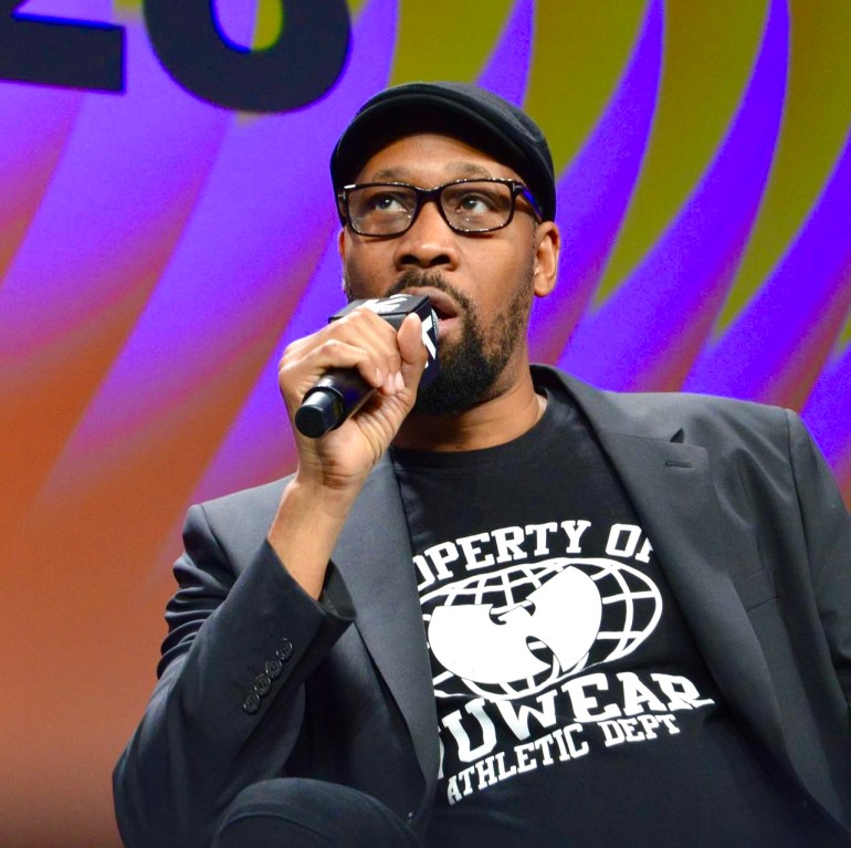 RZA can be seen glanced upwards while talking into a microphone. He is wearing all black. Black golf hat, black blazer, and a black “Property of Wuwear” shirt