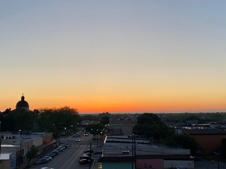 The top two thirds of the frame show an orange sky illuminating downtown San Marcus at dawn while the bottom third shows empty streets and buildings on the Square 