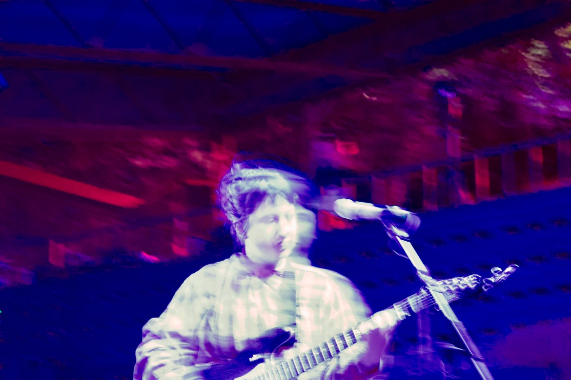 The photo shows Karly Hartzman of Wednesday performing on the outdoor stage at Mohawk. The photo is not fully in focus, giving it a wispy effect. The photo is a deep blue and pink color.