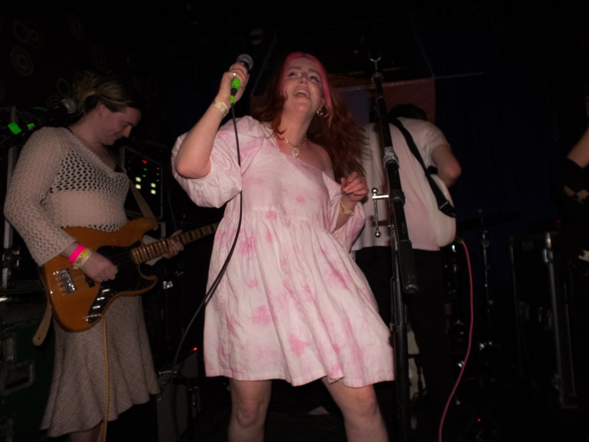 Jamie plays guitar in cream knitted sweater and skirt. Róisín (center) holds microphone wearing pink floral babydoll dress.