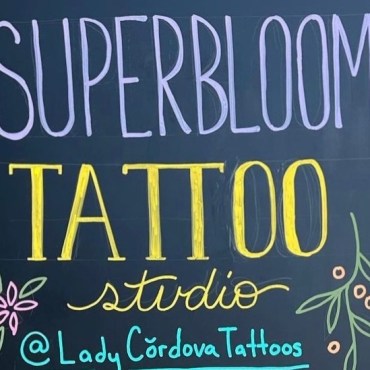 A cropped photo of a black chalkboard sign framed with light brown wood. The tect reads, ‘Super Bloom’ in large purple handwriting, followed by ‘Tattoo’ in yellow, ‘studio’ in small cursive yellow writing, and ‘@ LadyCordovaTattoos’ in teal. The words are framed by small drawings of various flowers.