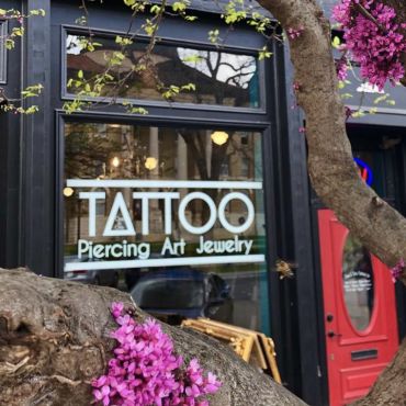 A picture of the exterior of One & Only Tattoo Shop, a black building with large windows and a red door. On the perimeters of the image, there is a tree blossoming with small pink flowers.