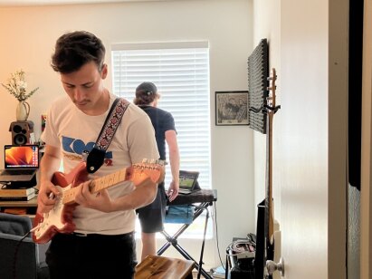 The band’s guitarist, Ray Chenevey, is playing guitar in a room with Conner Libera in the background watching a baseball game on an ipad.