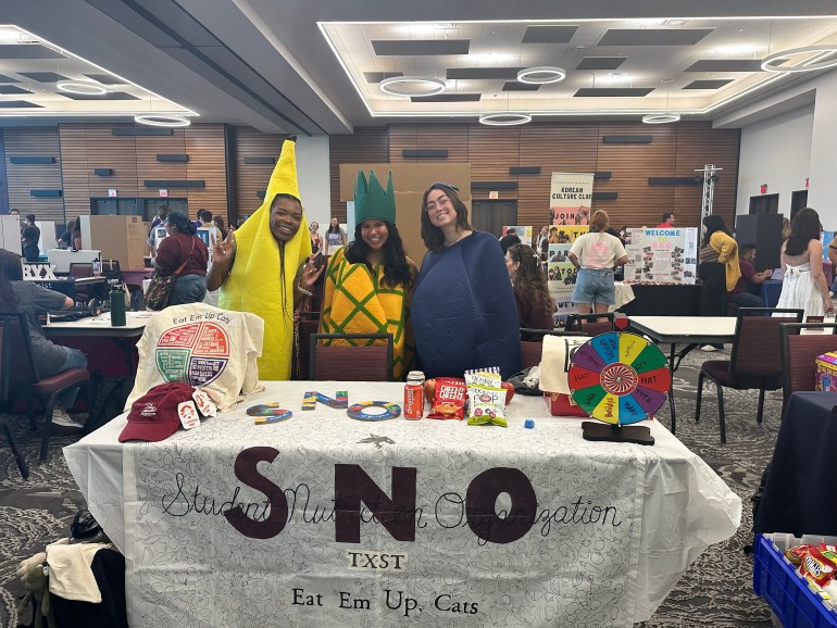 3 people dressed in fruit and vegetable costumes, standing behind Student Nutrition Organization table