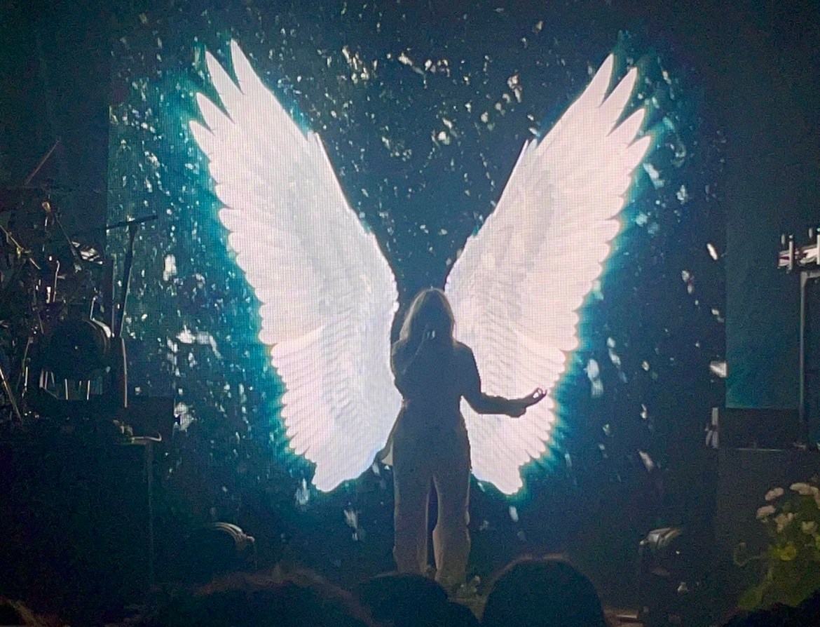 This image shows Reneé Rapp and her band performing the title track to her debut album Snow Angel for a crowd of people as angelic wings are positioned on a screen behind her.